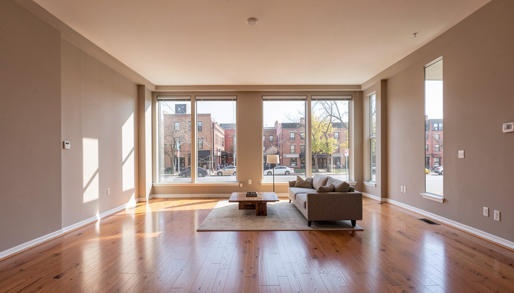 A sunlit interior of a spacious apartment living room with large windows, hardwood floors, and neutral-toned walls, overlooking a quiet DC street.