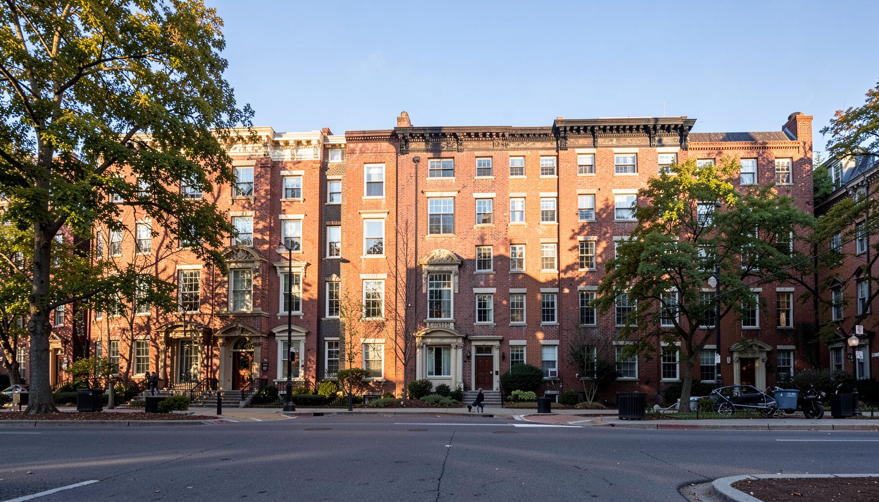 A row of classic brick rowhouses along a tree-lined Dupont Circle street on a clear day, with warm sunlight casting soft shadows across the facades.